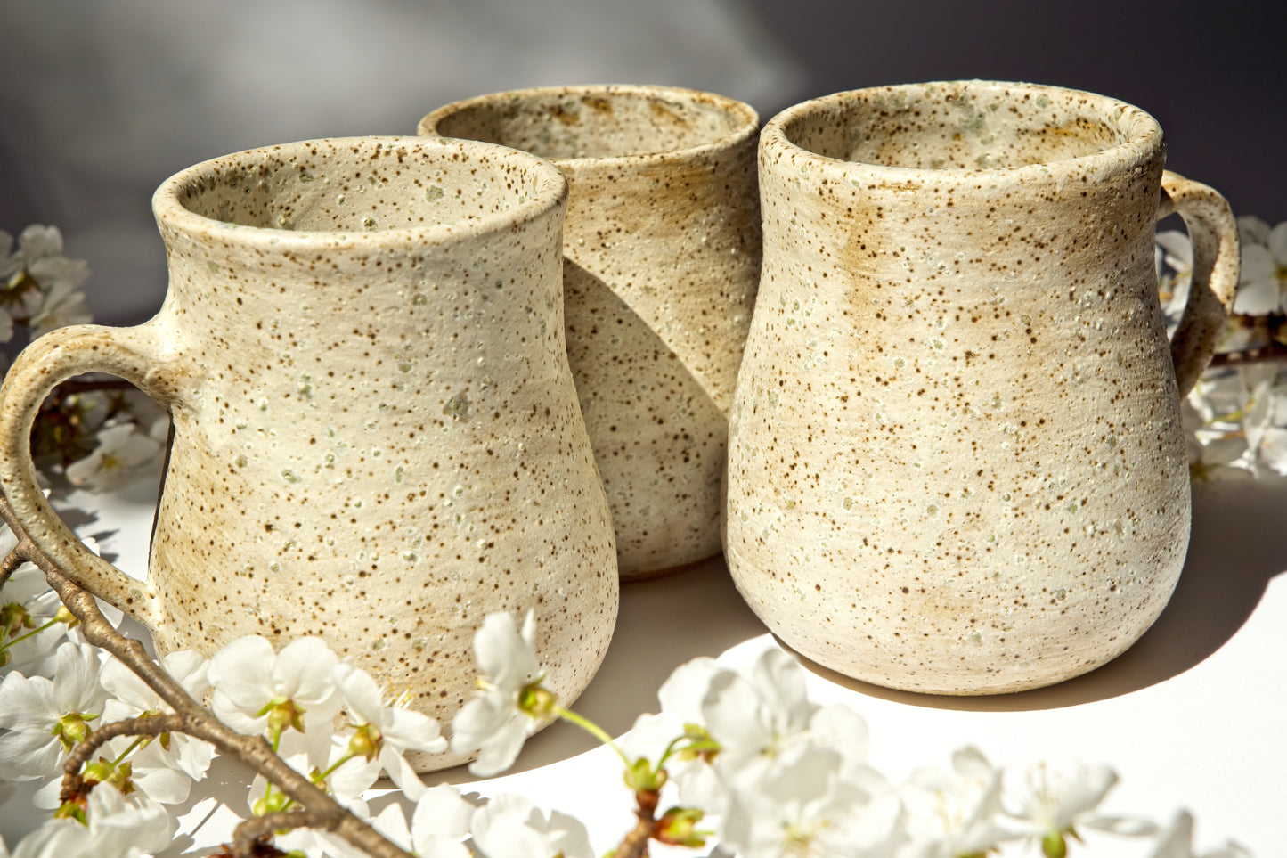 Three ceramic mugs with a speckled texture on a surface with white flowers.
