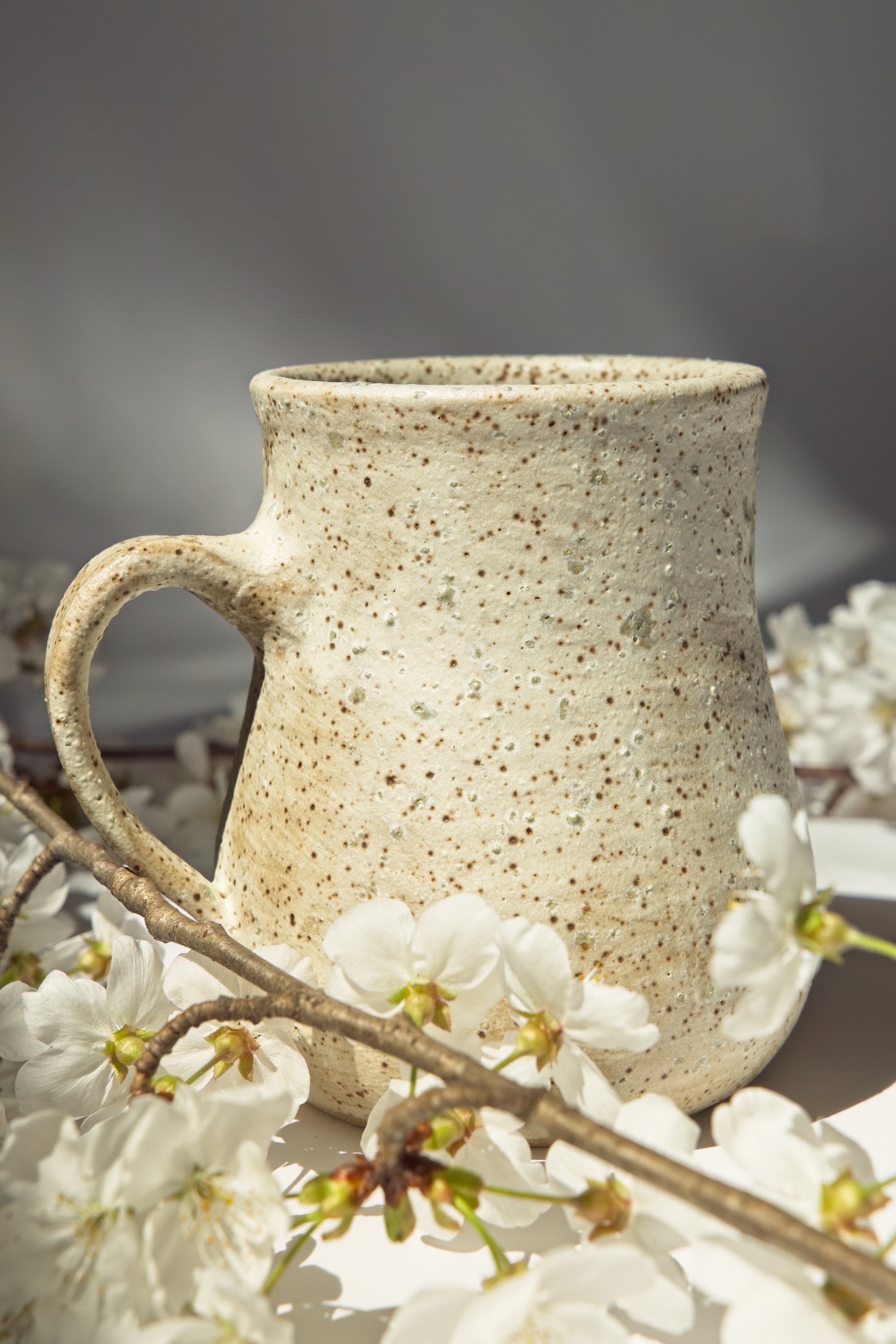 Speckled ceramic mug with a handle on a surface with white flowers and branches.