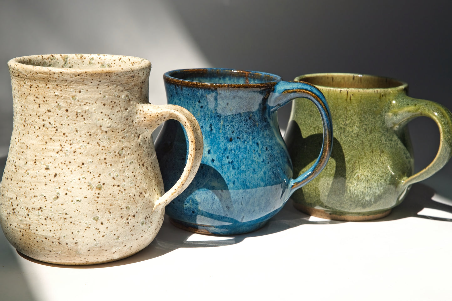 Three ceramic mugs in matte beige, blue, and green on a neutral background