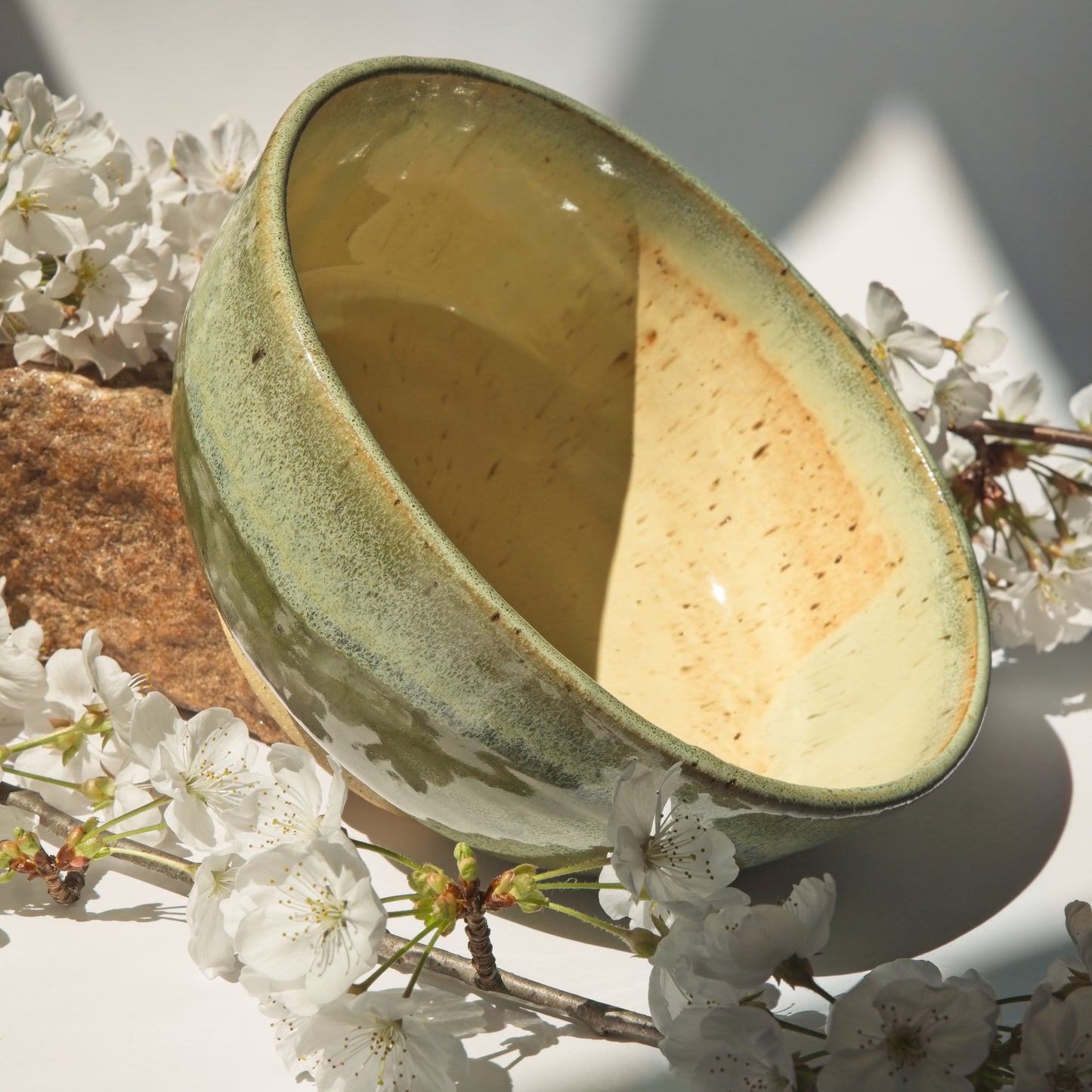 Large ceramic bowl with a textured green surface leaning against a rock and surrounded by cherry blossom branches