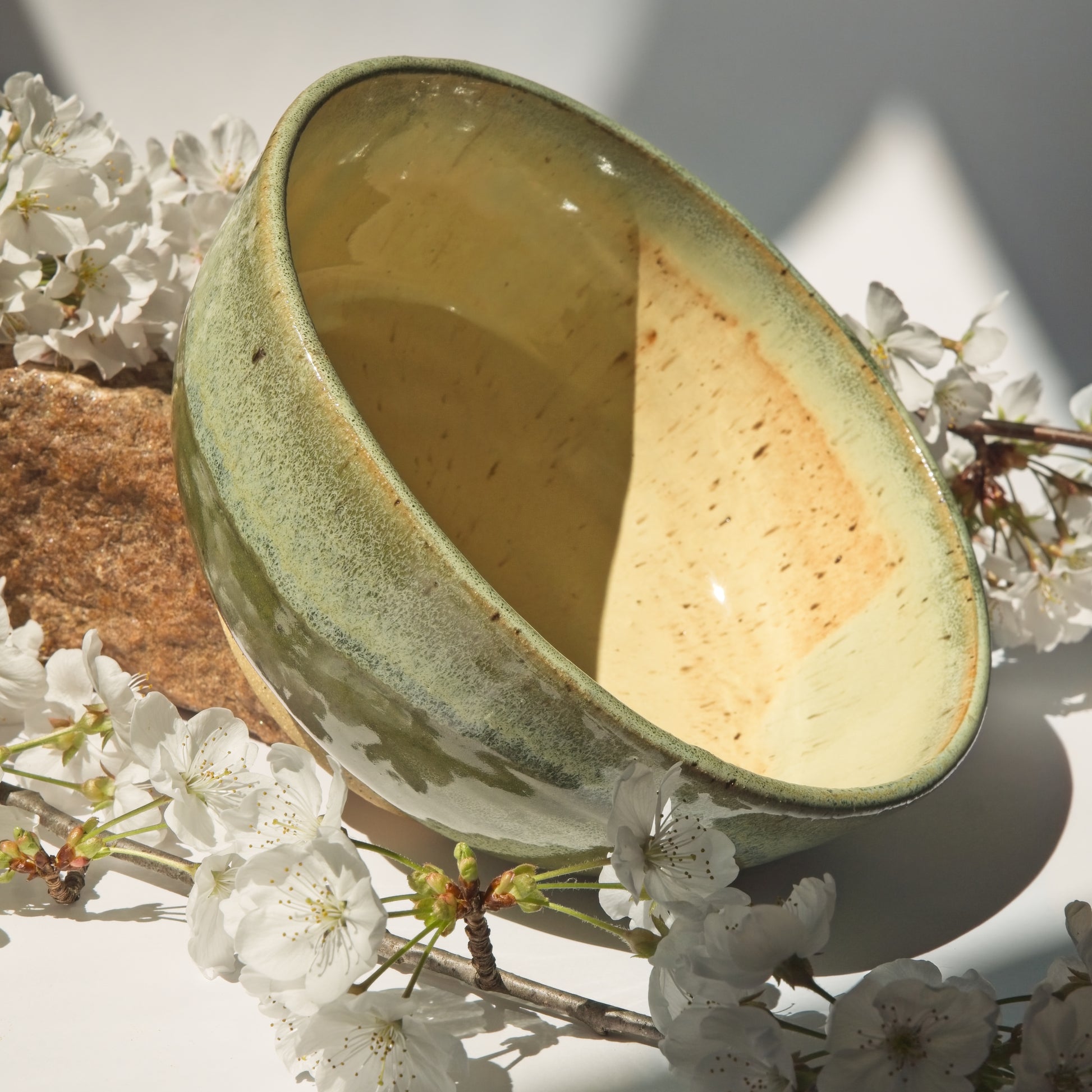 Large ceramic bowl with a textured green surface leaning against a rock and surrounded by cherry blossom branches