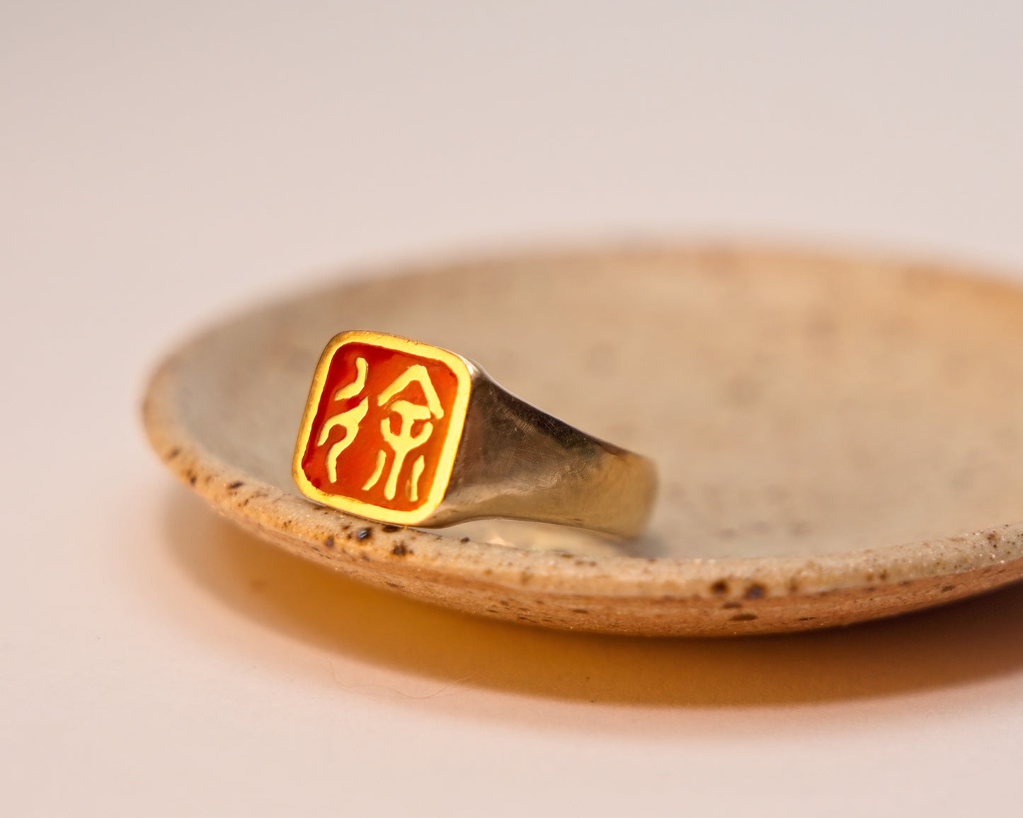 Gold ring with a chinese script within a red emblem on a matte ring dish with artistic speckles in the clay