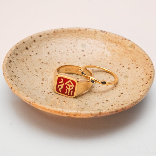 Gold rings, one with chinese characters in a red emblem, the other with small sapphires, sitting on a matte ring dish with speckled clay